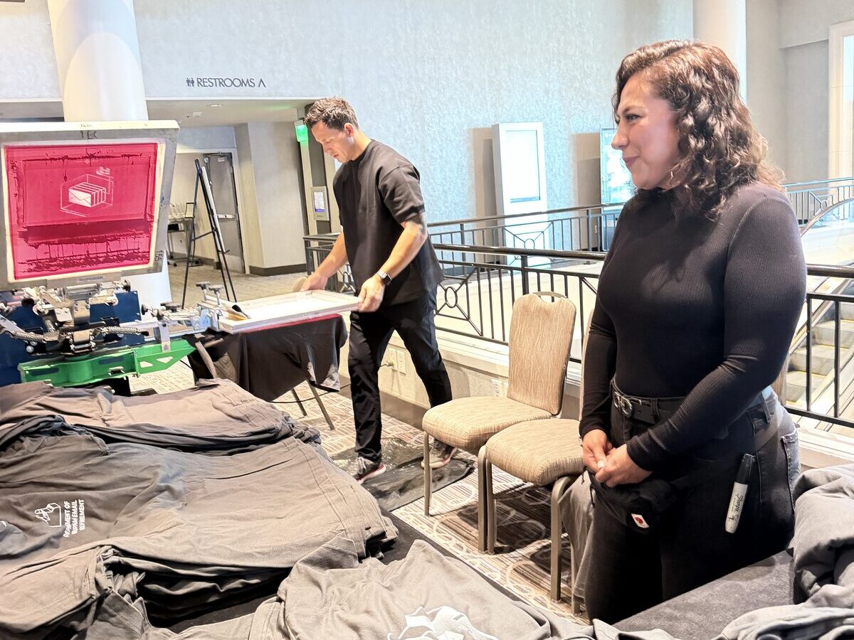 Two Merch Troop crew members at a press station beside tables of folded gray tees printed with a logo at a Denver corporate event