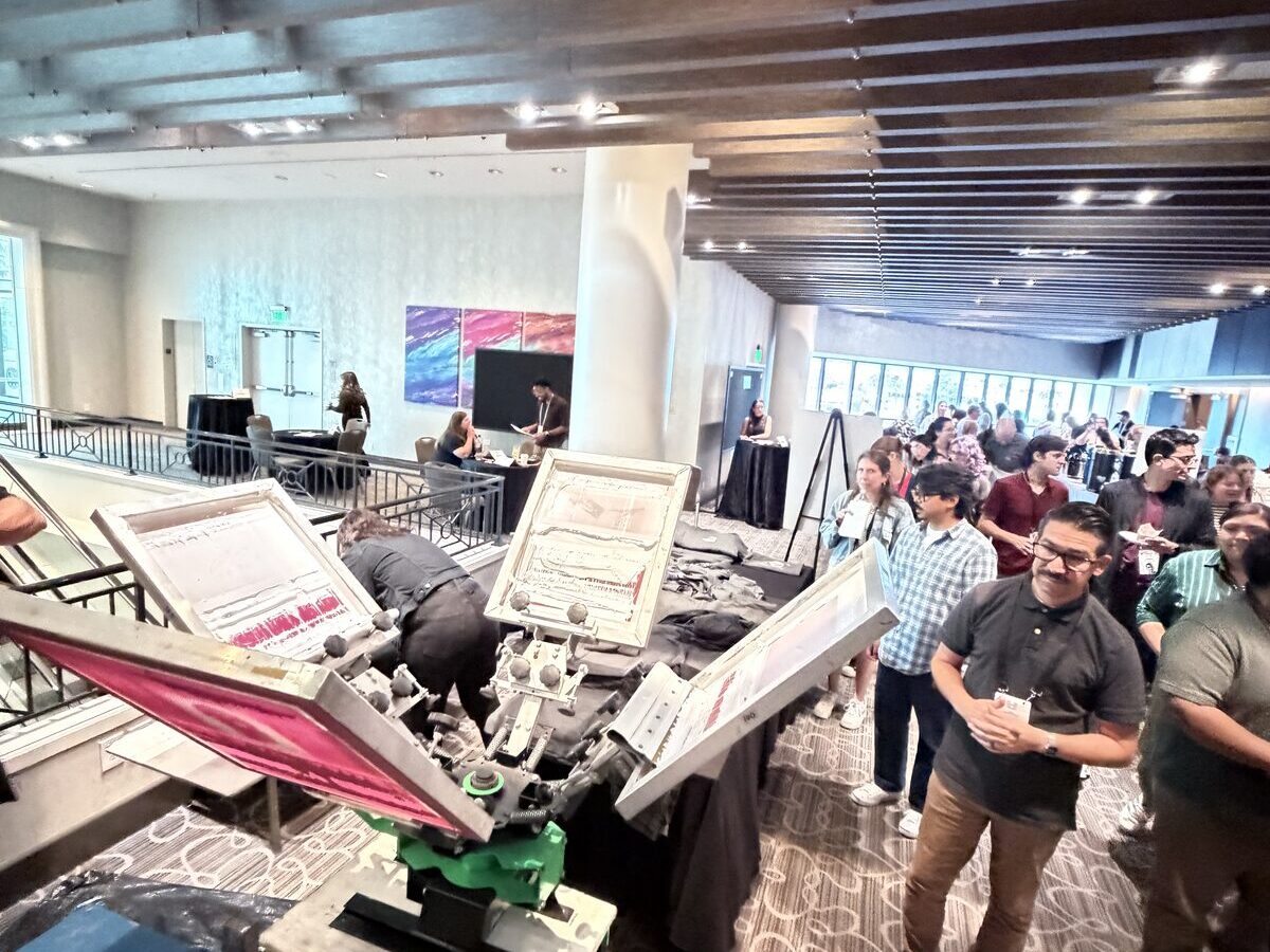 Guests lined up at a multi-color screen-printing carousel press inside a hotel ballroom at a trade-show-style event