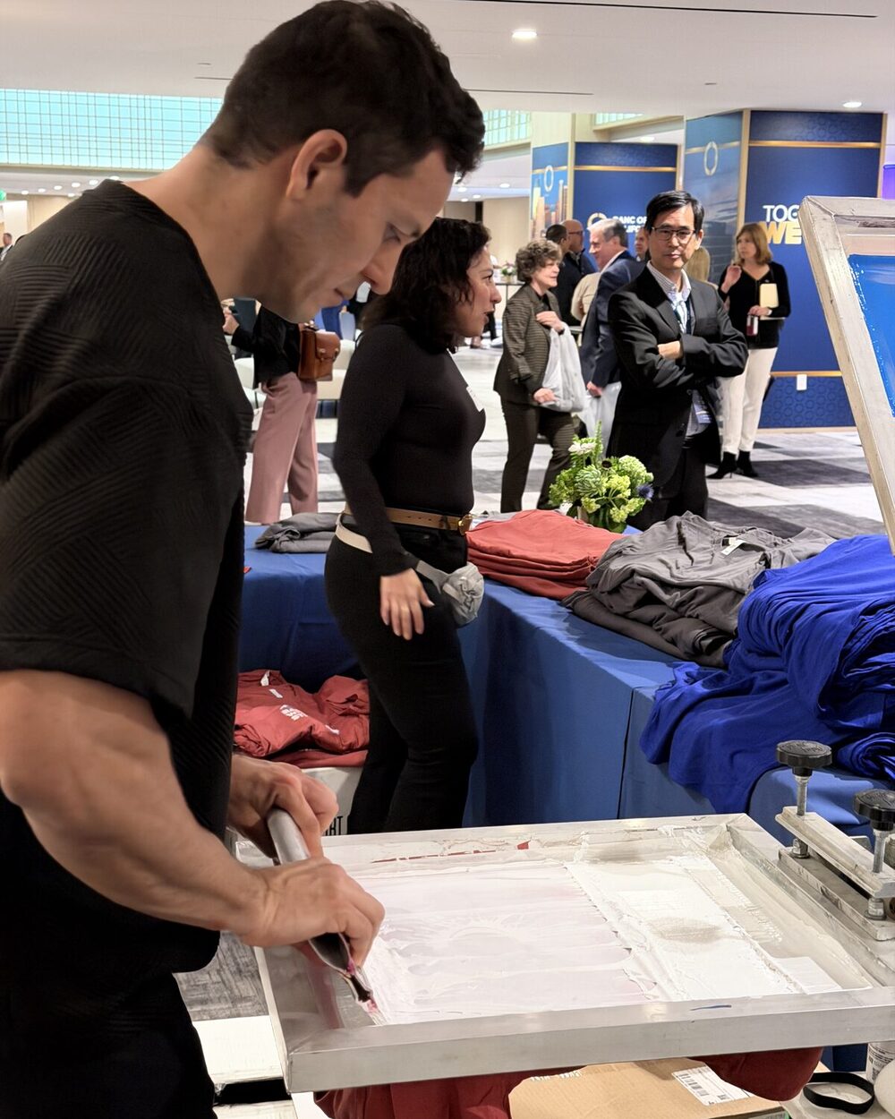 A Merch Troop printer pulling a squeegee across a screen on a manual press at a Denver-area event while guests look on near a table of folded tees