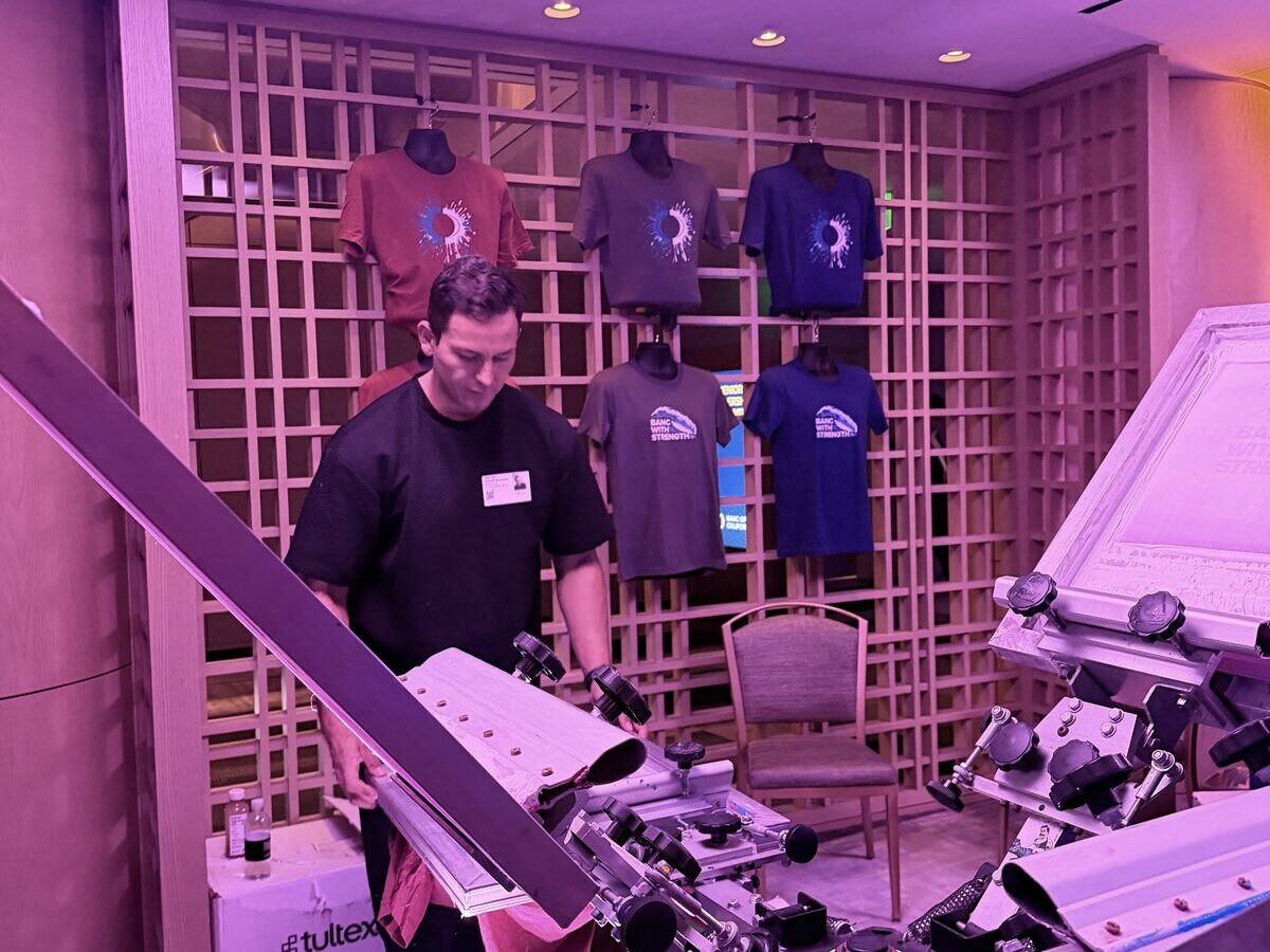 A screen-printing carousel press with a wall of freshly printed event tees on display behind it under colored lighting
