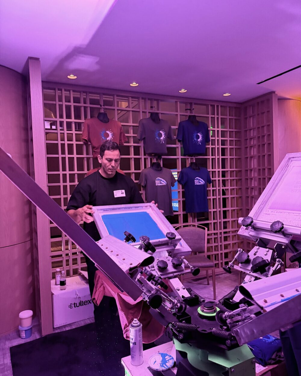 A Merch Troop printer holding a screen frame in front of a wall display of freshly printed event tees under colored lighting